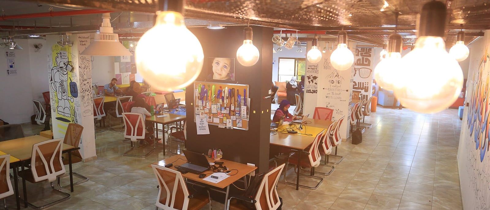 Großraumbüro mit Glühbirnen-Lampen, Arbeitstischen und Laptops, im Hintergrund arbeiten einige Menschen
