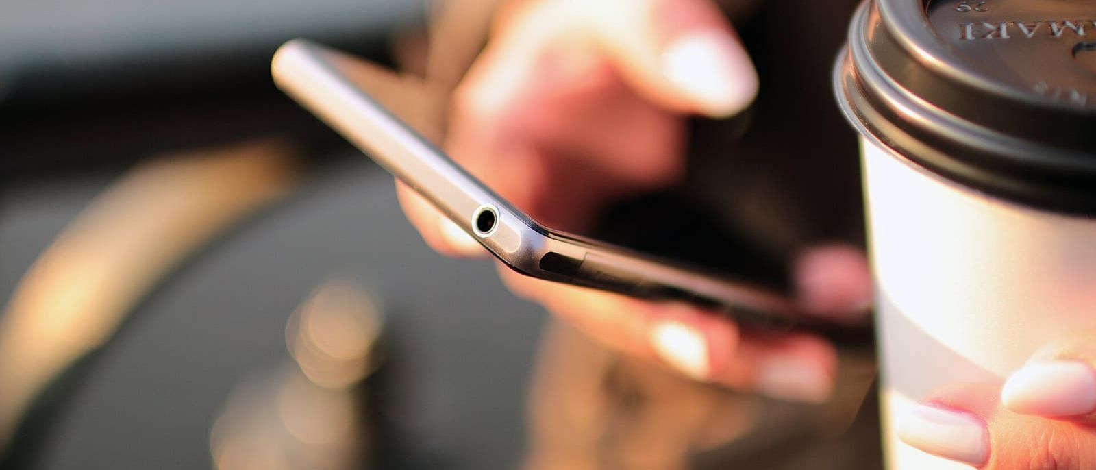 Hand hält ein Smartphone, daneben ein Kaffeebecher zum Mitnehmen im Gegenlicht