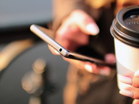 Hand hält ein Smartphone, daneben ein Kaffeebecher zum Mitnehmen im Gegenlicht