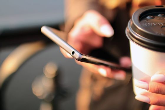 Hand hält ein Smartphone, daneben ein Kaffeebecher zum Mitnehmen im Gegenlicht