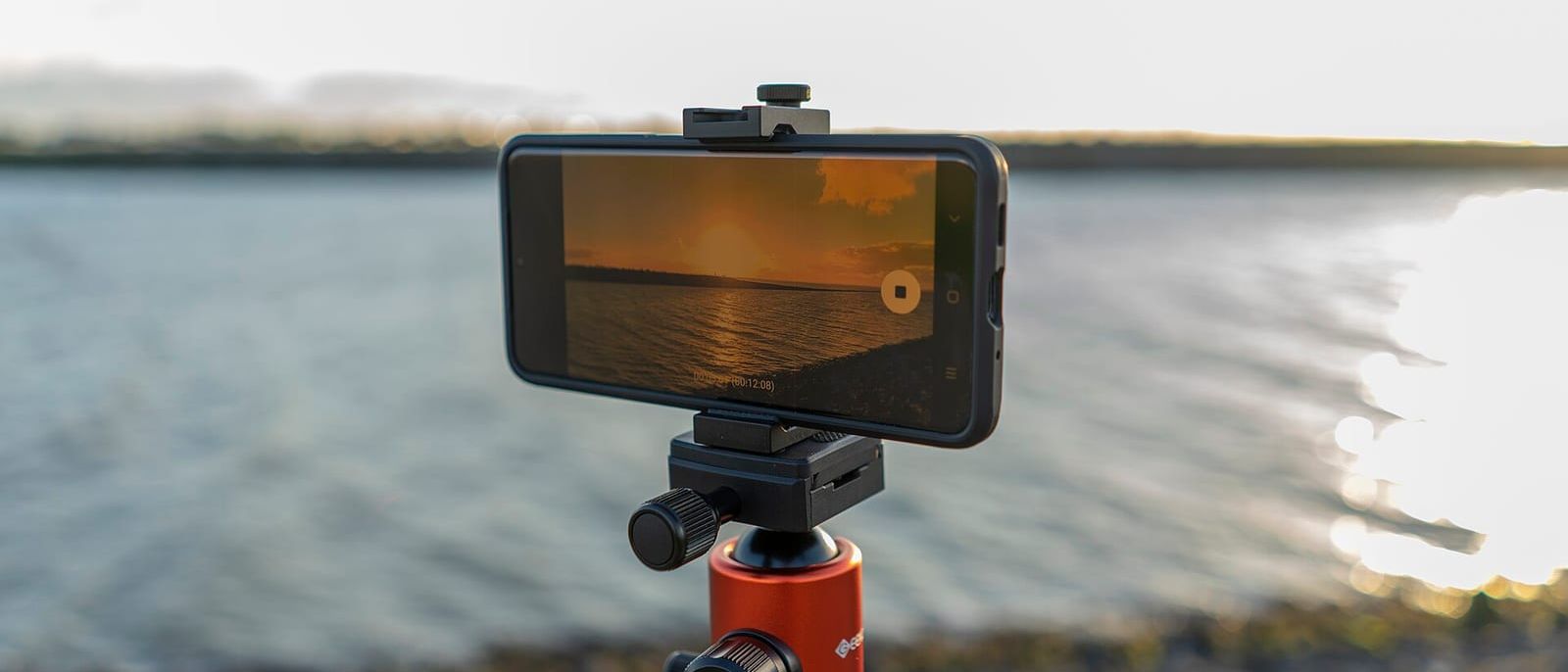 Smartphone auf einem Stativ filmt einen Sonnenuntergang am Wasser, Display im Fokus