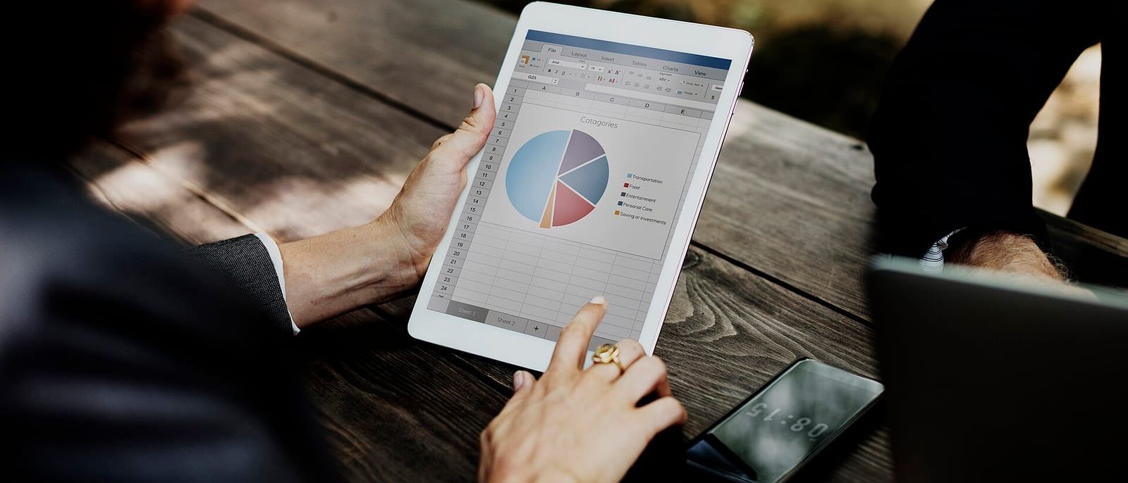 Hände halten ein Tablet mit Tabelle und Kreisdiagramm über einem Holztisch, daneben ein Smartphone